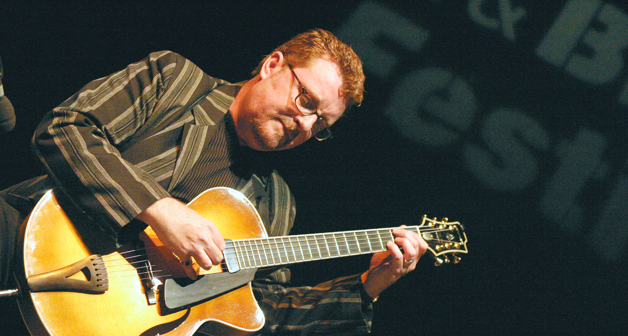 Martin Tyler plays a Jazz arch-top onstage. He is wearing a striped blazer (a little like a stockbroker) and fretting a chord.